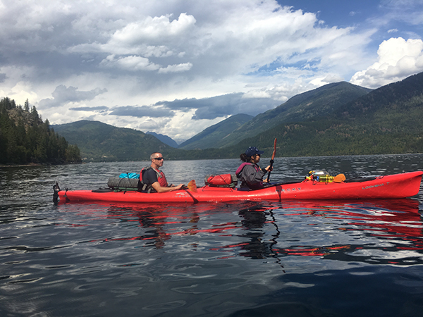 Two people paddle a red tandem kayak across calm lake water. The kayak carries gear secured on deck, with forested mountains rising on both sides and dramatic clouds filling the sky above.