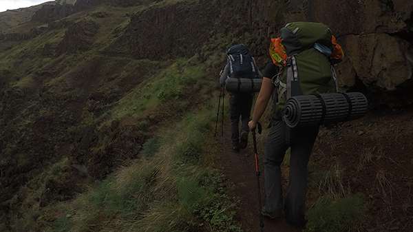 Hikers on an outdoor path trek towards a mountain peak surrounded by greenery. They wear full backpacks with gear for camping attached.