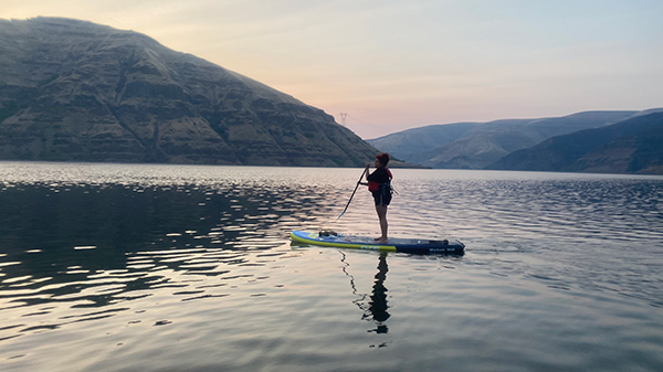 A person stands up paddleboarding on calm water at sunset. The paddleboard glides across the lake as the sky reflects soft pink and blue hues, with rolling hills and mountains rising along the shoreline in the background.