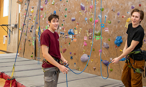 Two people stand on the padded floor of an indoor climbing gym, holding blue climbing ropes and wearing harnesses. Behind them is a tall climbing wall covered with colorful handholds, ropes hanging from above.
