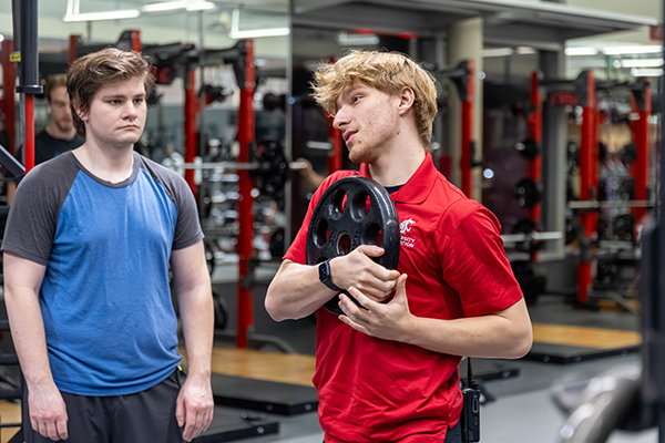 Inside a weight training facility, two people stand near squat racks and gym equipment. One person in a red shirt denoting a Personal Trainer demonstrates a strength or fitness concept while holding a weight plate, and another person stands nearby listening. Free weights, racks, and mirrors fill the background of the gym.