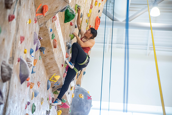 A person wearing a harness climbs an indoor rock wall, reaching for a green handhold among colorful climbing holds. A safety rope is attached above, with gym lighting and structural beams visible in the background.