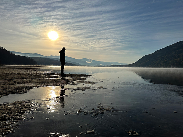 A single person stands near the edge of a partially frozen lake at sunrise, silhouetted against a low sun. Snowy mountains and forested hills surround the calm water, which reflects the sky and figure, creating a quiet winter landscape.