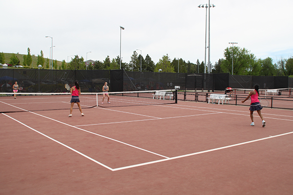 Four people play a doubles tennis match on outdoor campus courts, with players positioned on both sides of the net. Multiple tennis courts, benches, light poles, and trees are visible in the background under an overcast sky.