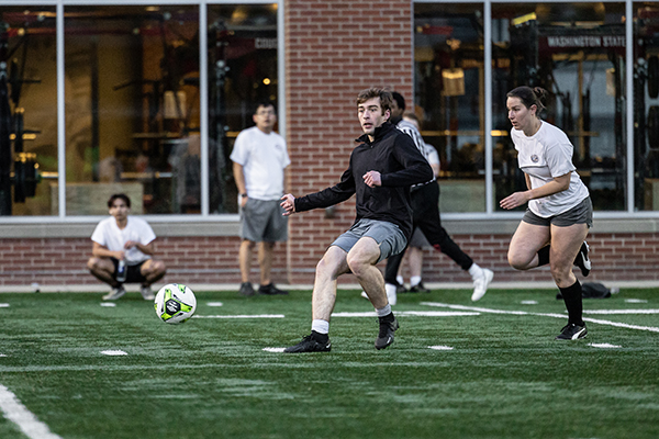 Several people play outdoor soccer on a turf field, with one person in the foreground kicking a green-and-white soccer ball while others run nearby to challenge the play. A brick building with large windows and additional people watching appears in the background.