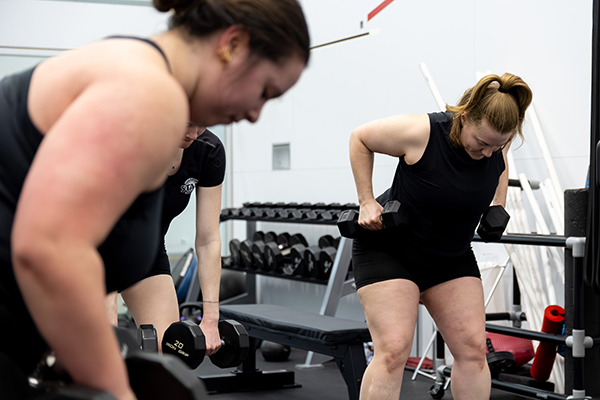 In a fitness weight room, two participants perform dumbbell exercises near a bench and a rack of free weights. One participant is bent forward lifting dumbbells while another stands nearby holding weights; additional gym equipment and mirrors are visible in the background.