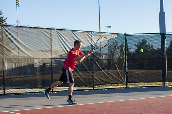 A person hits a tennis ball with a racket during a singles match on an outdoor campus tennis court, with a chain-link fence, light poles, and clear sky in the background.