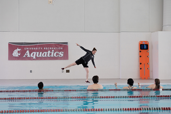 An aqua aerobics class takes place in an indoor pool. An instructor stands on the pool deck demonstrating a balance movement while several participants follow along from the water; lane lines cross the pool in the foreground, and a wall banner reads “University Recreation Aquatics” near an orange rescue backboard.